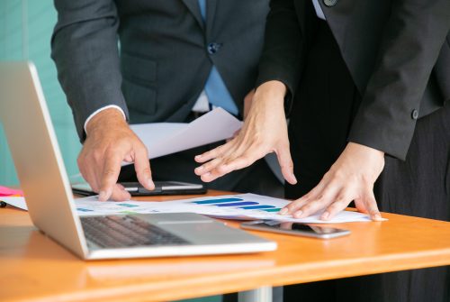 Unrecognizable businesspeople studying statistics and holding papers with hands. Office employees standing near table with laptop. Man pointing with finger. Business, teamwork and management concept